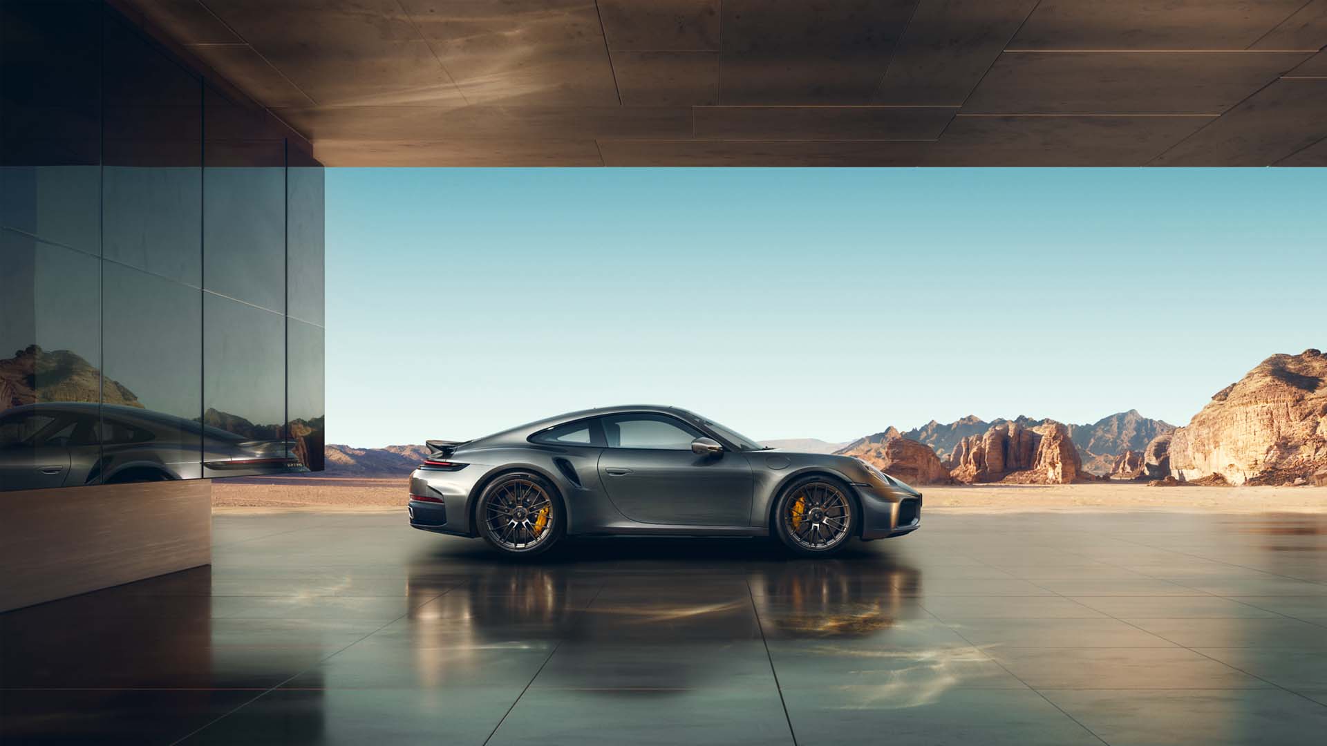 Side profile of a gray Porsche 911 Turbo S parked in a modern architectural covered space with polished reflective floor, overlooking a vast desert landscape with rocky mountains under clear blue sky